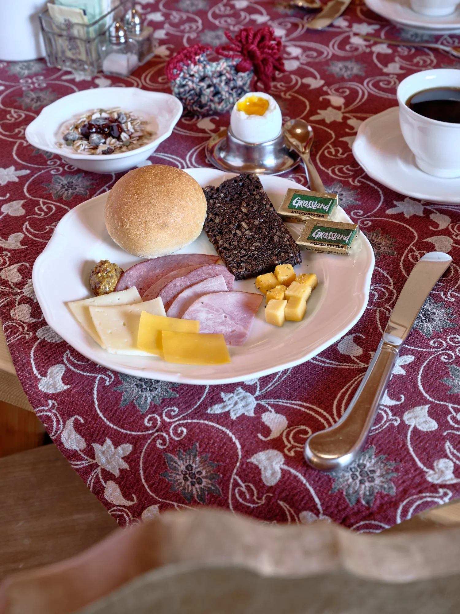 A plated Pension Anna breakfast — fresh roll, dark rye, meats, cheese, soft-boiled egg, and coffee