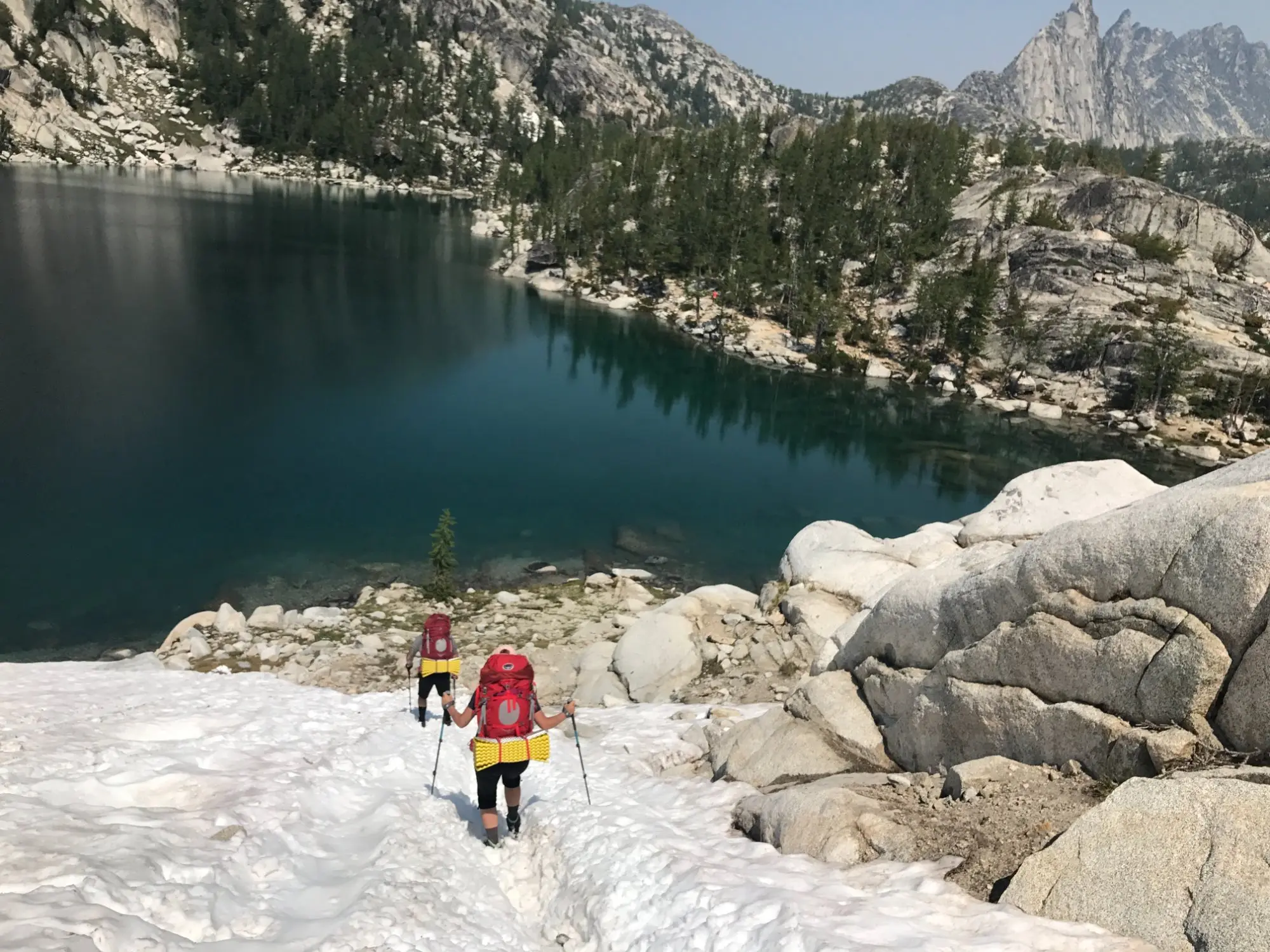 Two hikers descending a snowfield to a turquoise alpine lake in the Enchantments